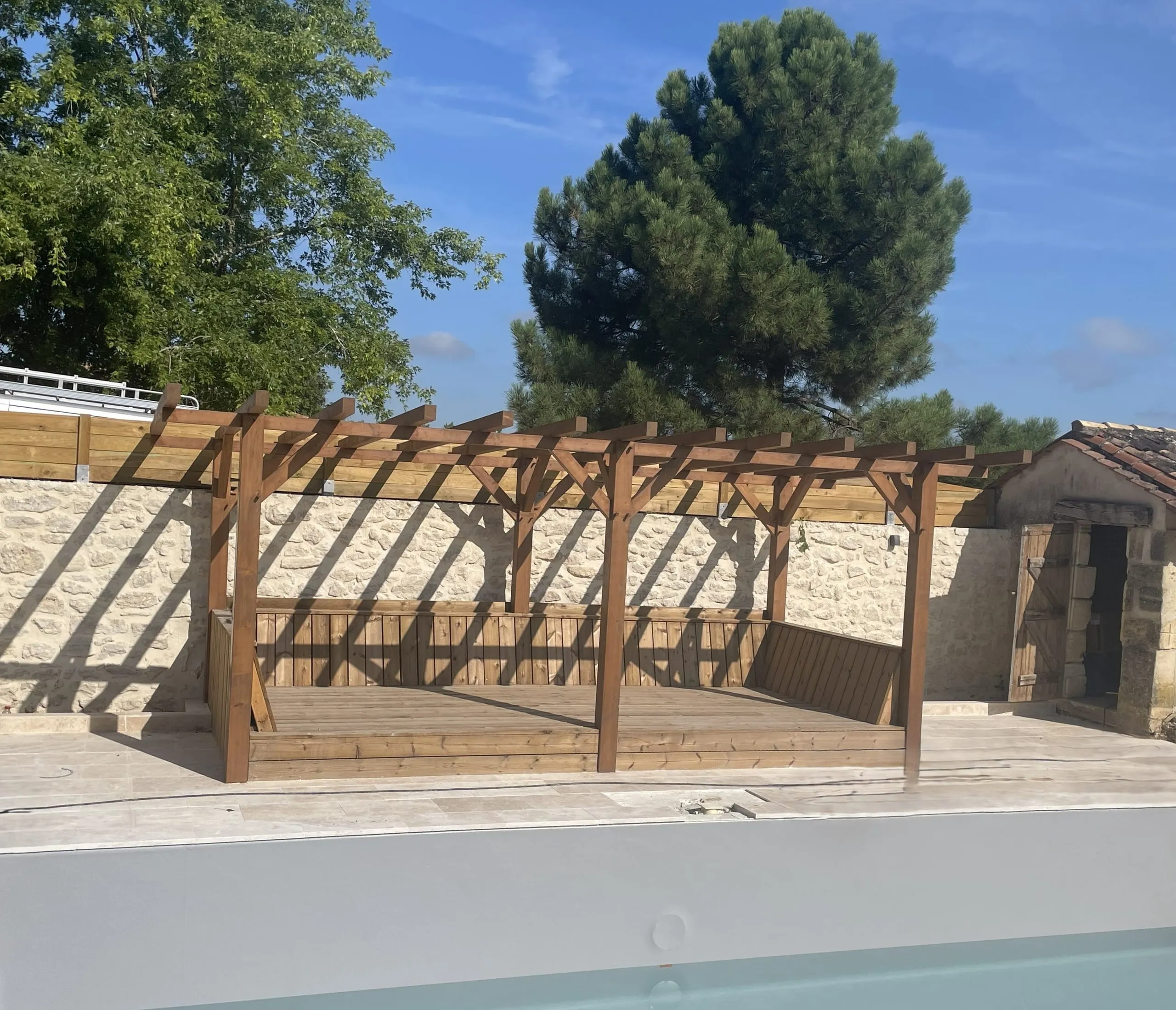 Pergola en bois avec banquettes intégrées et terrasse aménagée devant un mur en pierre