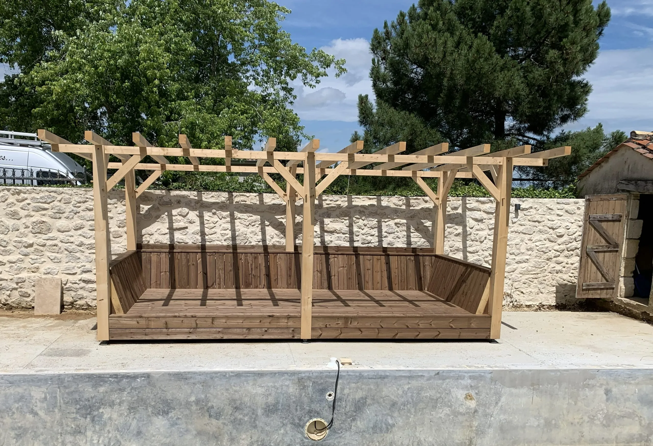 Pergola en bois clair avec banquette intégrée devant un mur en pierre