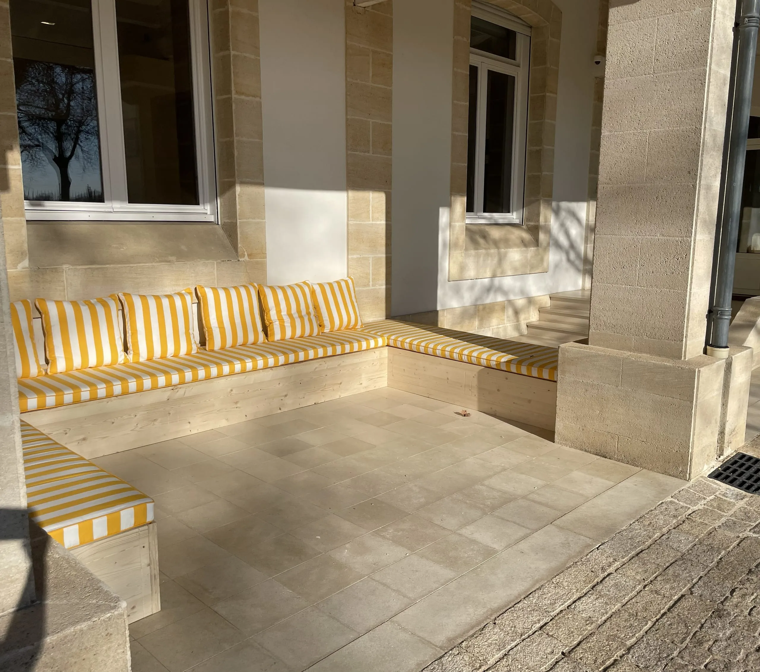 Banquette d’angle en bois clair avec coussins rayés jaunes et blancs sous une terrasse couverte