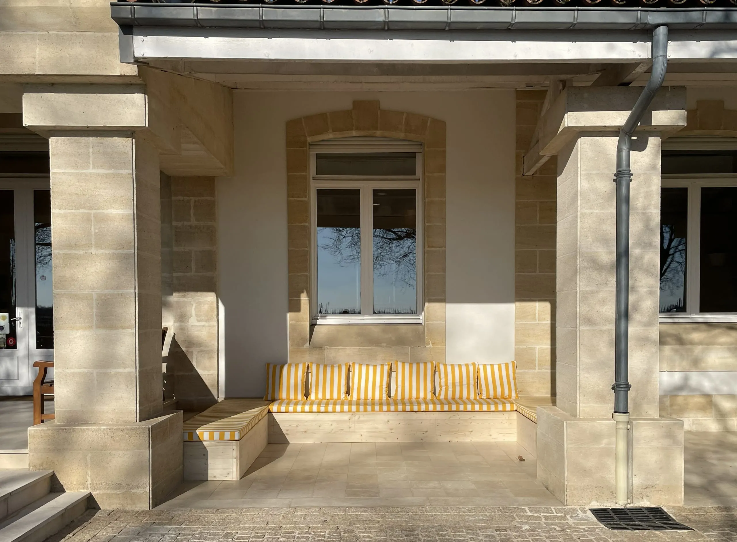 Banquette extérieure en bois avec coussins jaunes et blancs devant un mur en pierre